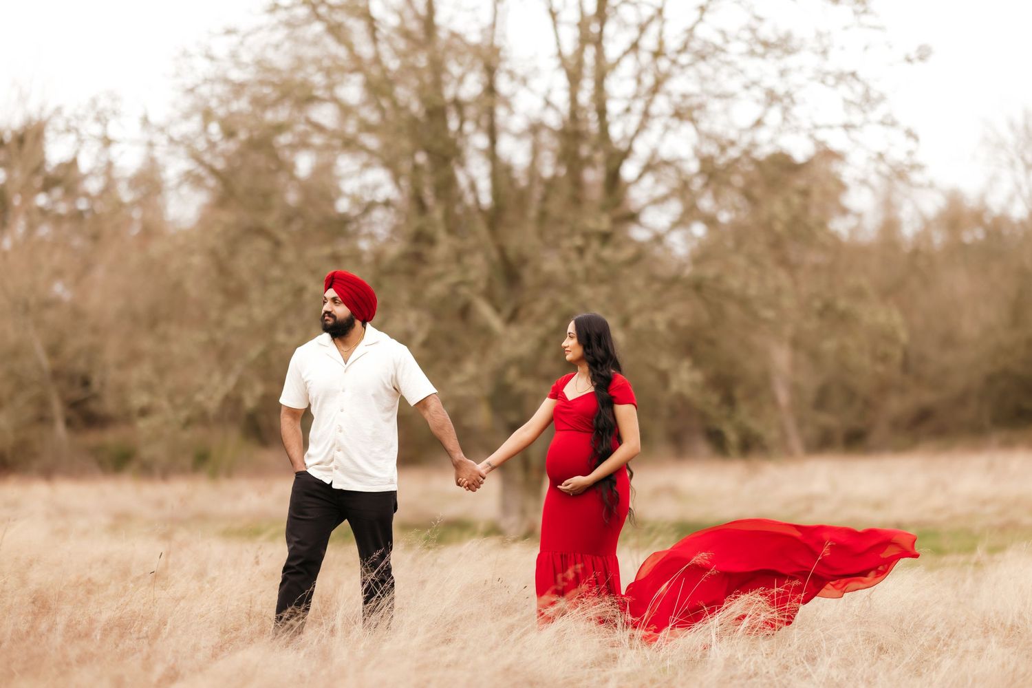 A couple holds hands in a field with a red dress flowing dramatically in the breeze.