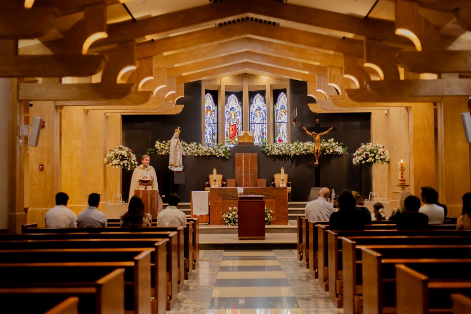 Catholic church interior with wooden pews, stained glass windows, and floral altar arrangements.
