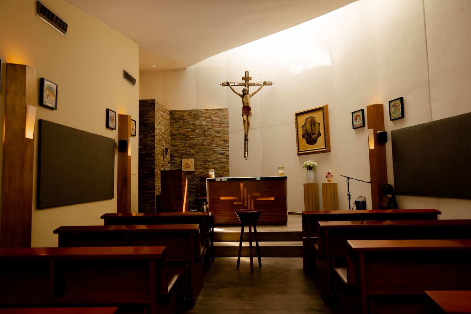 Small chapel interior with wooden pews, crucifix, religious icons, and stone wall accent.