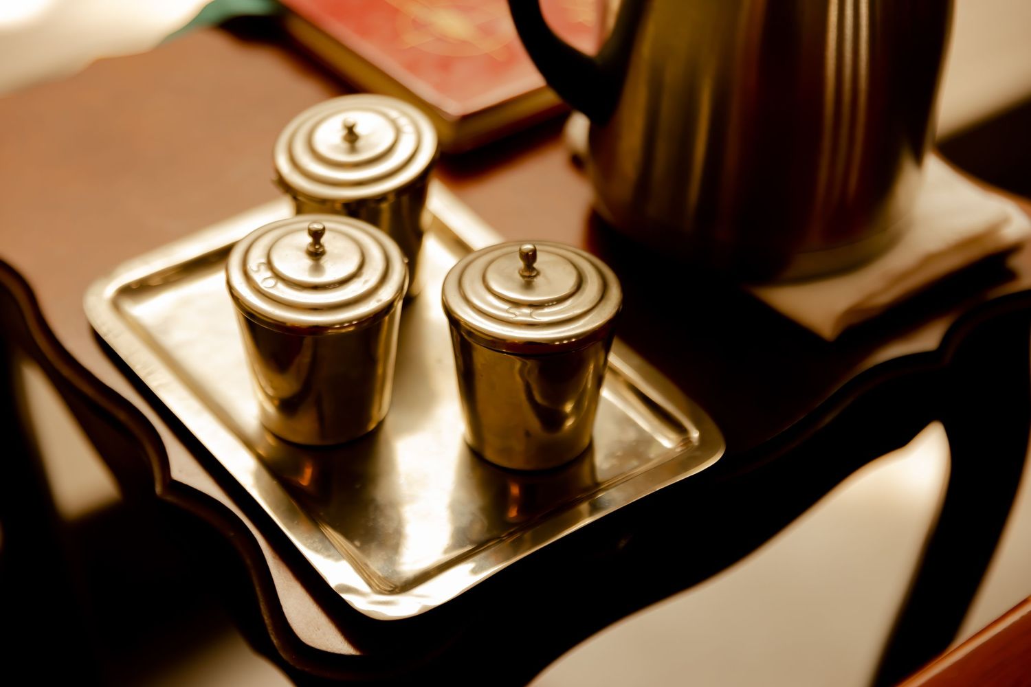 Vintage silver holy oil containers on a tray beside a black teapot in warm light.