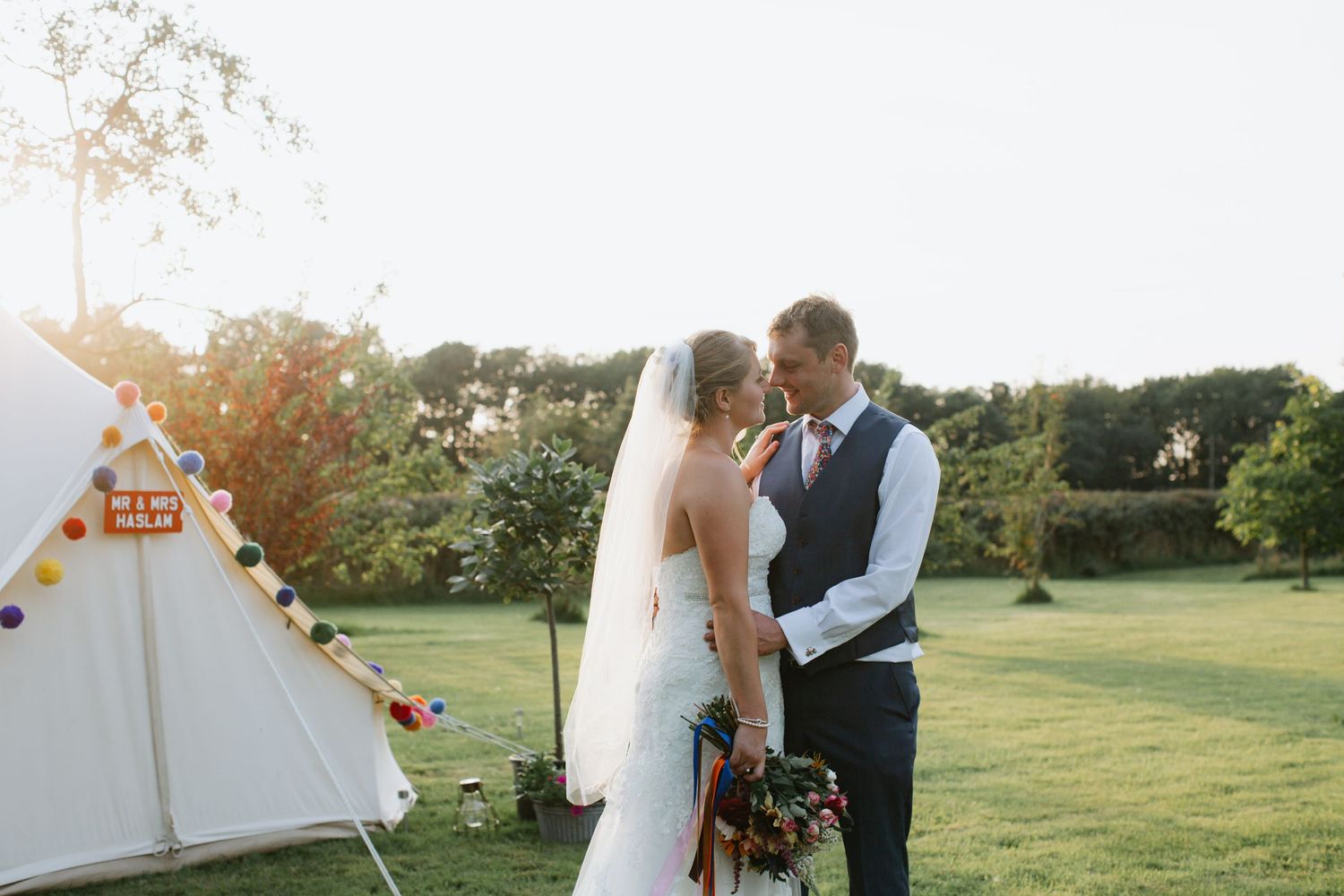 A sunset wedding moment with a couple standing near a tipi tent in a garden setting.
