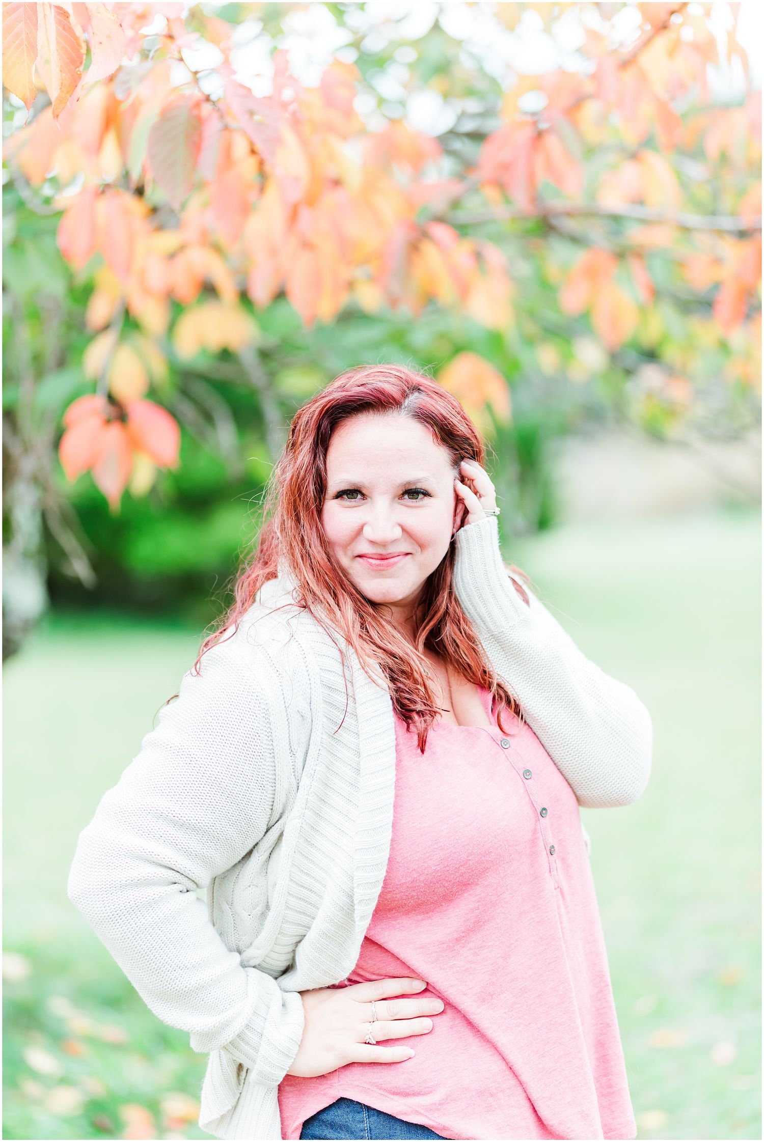 Person in white sweater and pink top poses outdoors with autumn foliage in background.