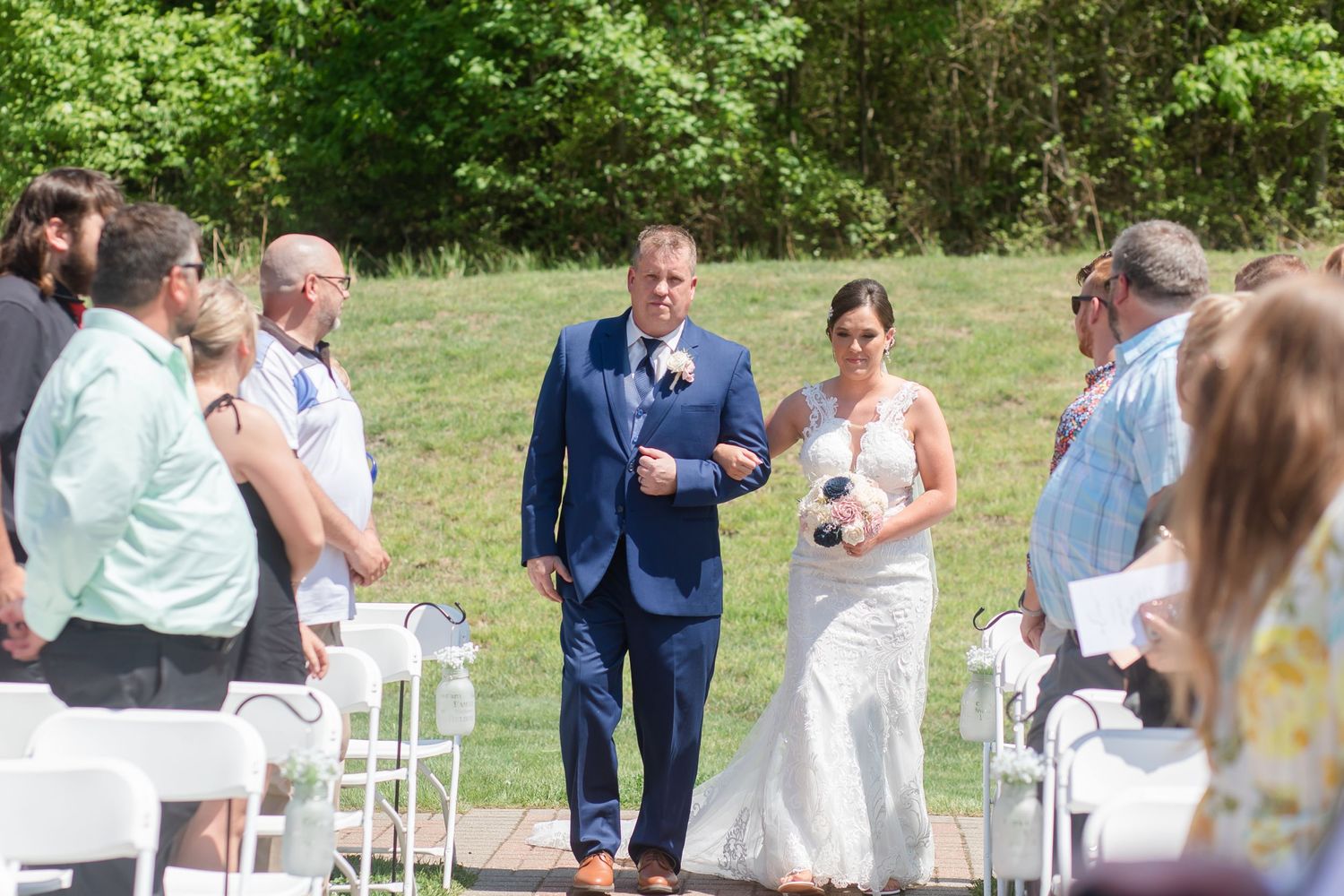 Wedding guests gather outdoors for a celebration under a rustic wooden pergola on a sunny day.