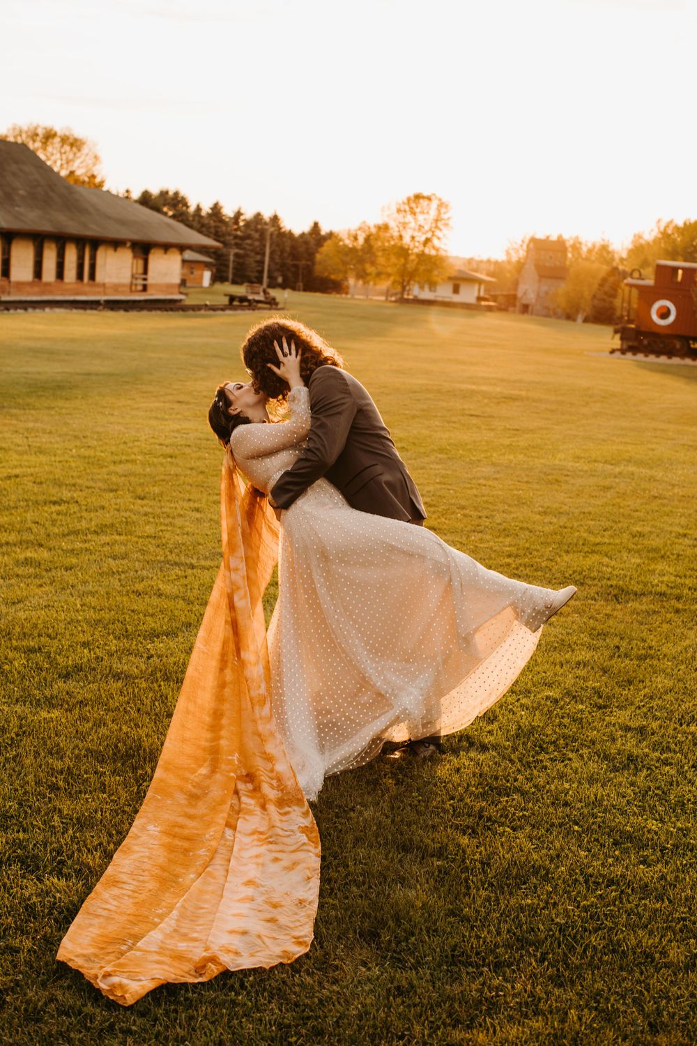Groom lifts bride in romantic dip kiss on green lawn at golden sunset with orange flowing veil.