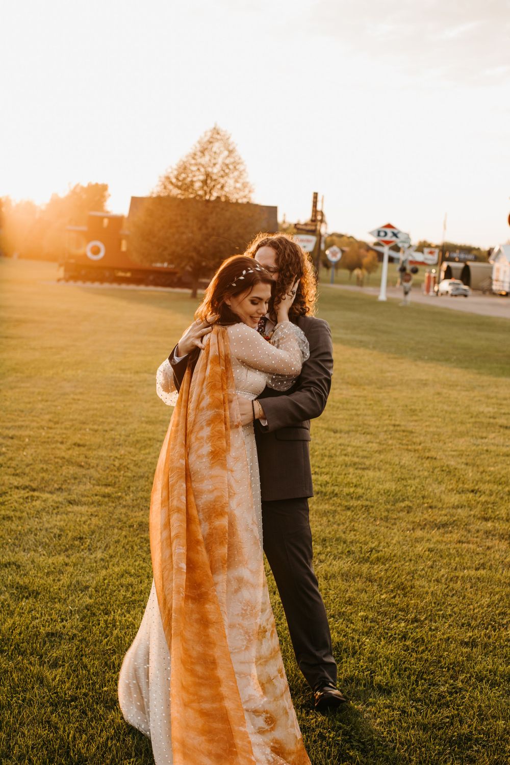 Groom embraces bride from behind on sunlit lawn as her orange tie-dye veil flows in the breeze.