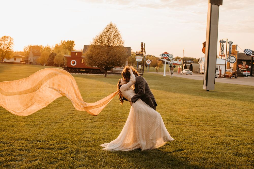Groom dips bride in passionate kiss on green lawn as her long orange veil billows in the wind.