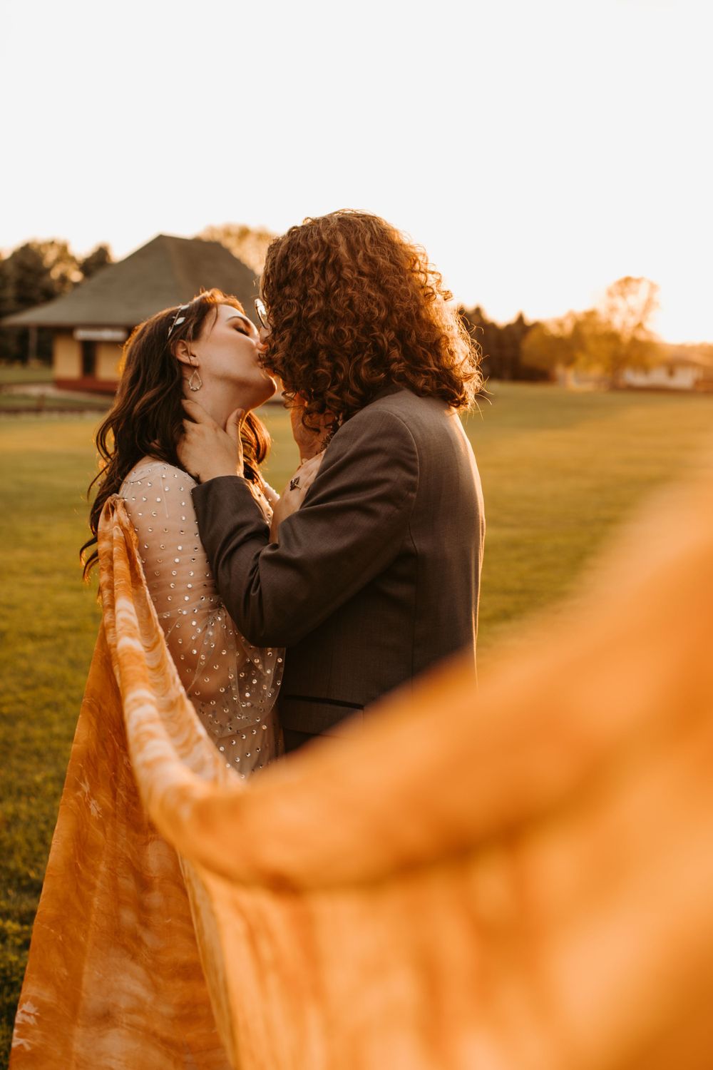 Couple shares a kiss at golden hour as brides orange veil flows beautifully in the foreground.
