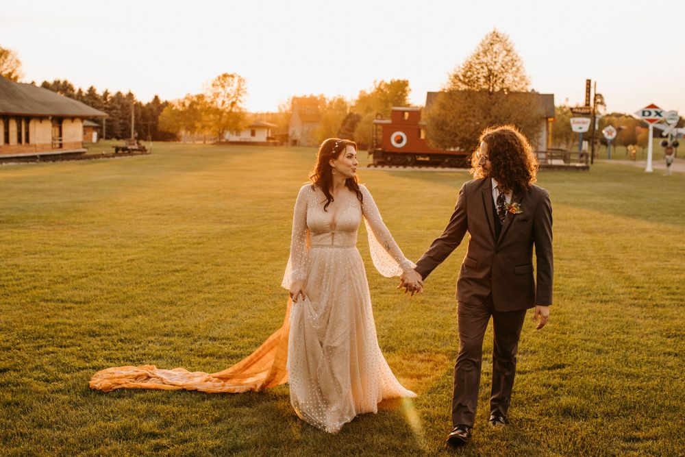 Bride and groom hold hands walking across sunlit green lawn with orange veil trailing behind her.