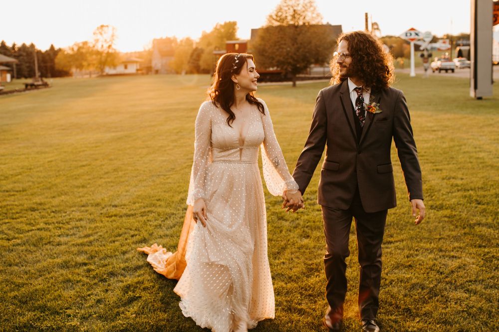 Bride and groom smile at each other while holding hands and walking through golden sunset light.