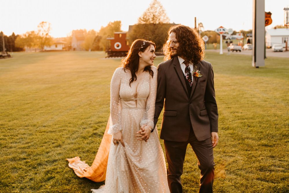 Bride and groom gaze lovingly at each other holding hands on green lawn during golden hour sunset.