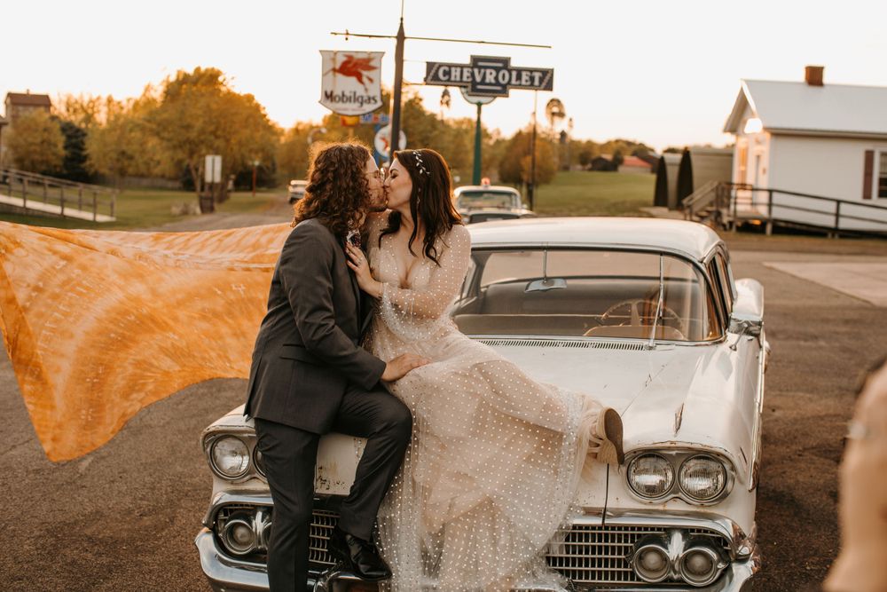 Bride and groom kiss passionately on a vintage white Chevrolet car at sunset during their wedding.