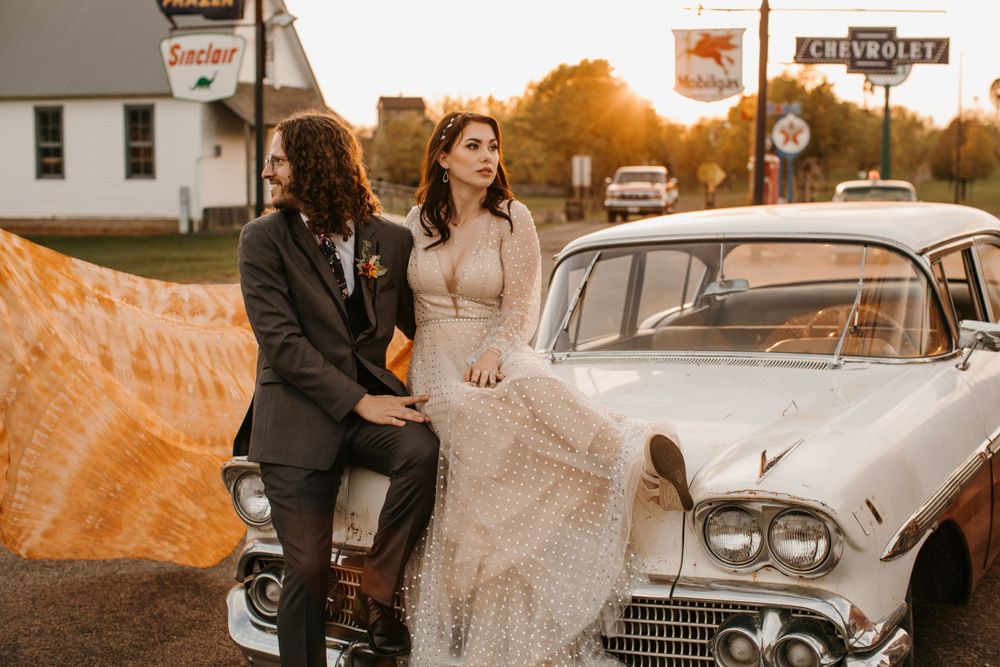 Bride and groom pose beside a classic vintage white Chevrolet car with an orange veil flowing in the breeze.