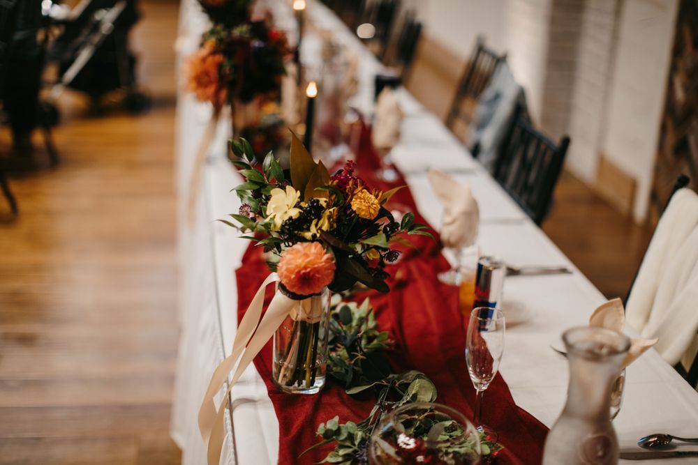 Long wedding reception table decorated with a red runner, colorful fall floral arrangements, and ribbon accents.