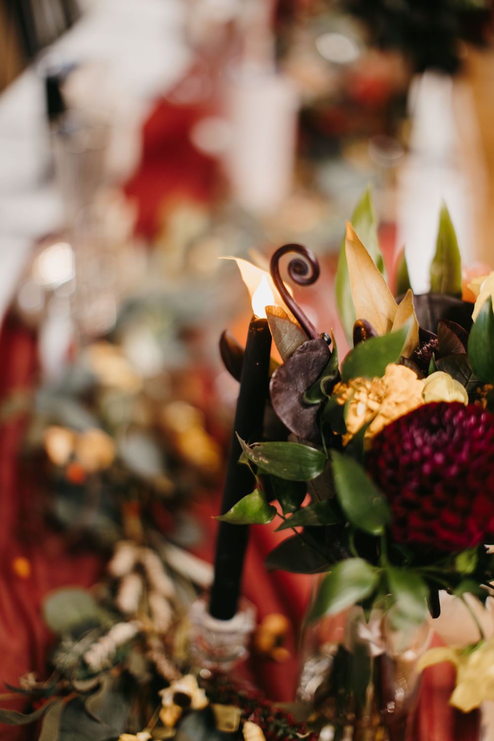 Elegant fall wedding centerpiece with burgundy dahlias, orange roses, dark calla lilies, and black taper candles.