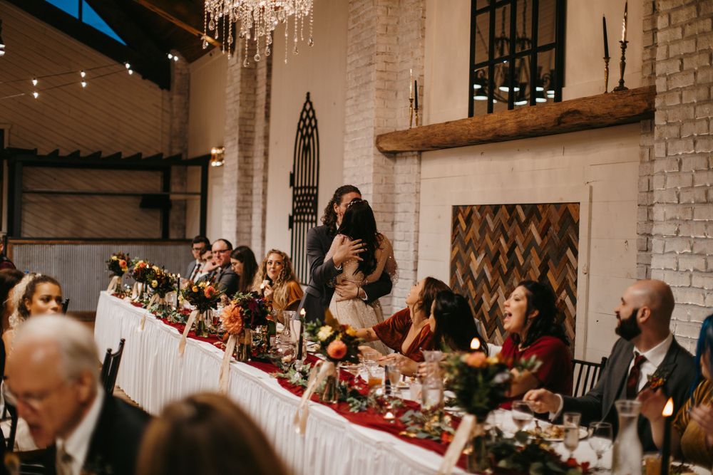 Wedding reception guests seated at a long decorated head table with red linens and colorful floral centerpieces.