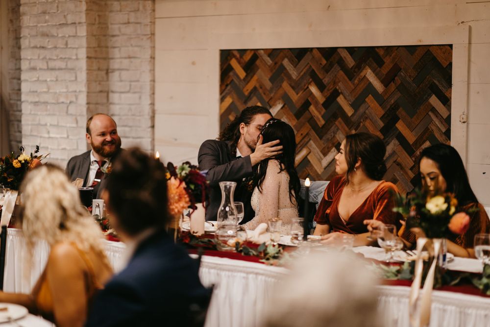 Bride and groom sharing a tender embrace at the wedding reception head table with wood herringbone backdrop.
