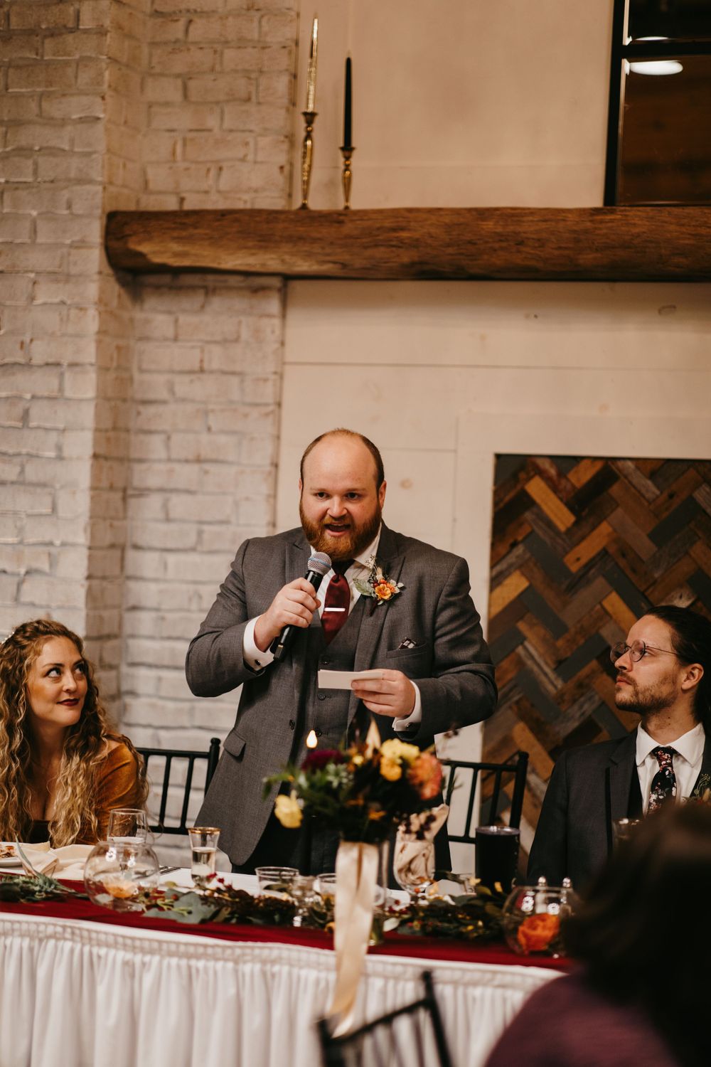 Best man giving a wedding toast at the head table in a rustic white brick venue with wood accents.