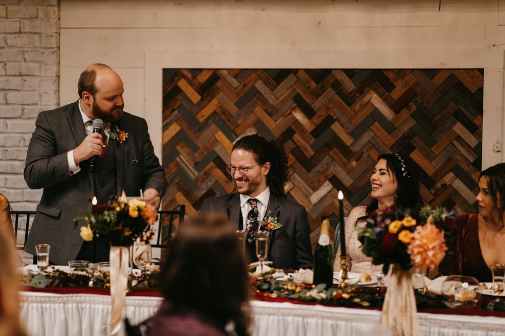 Groomsman delivering a heartfelt wedding toast as the smiling groom and bride listen at the reception table.