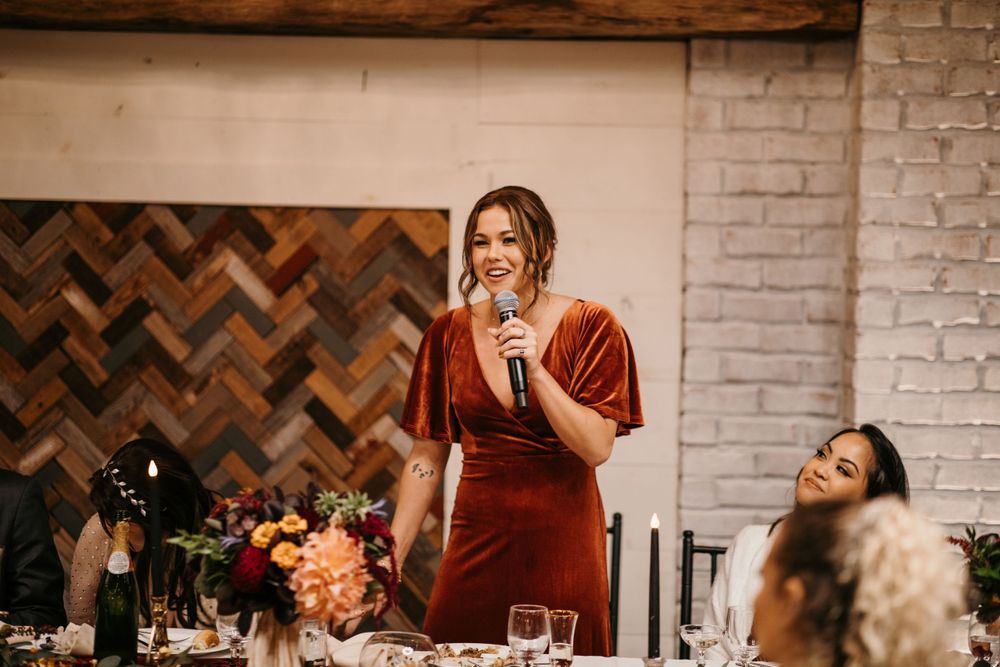 Woman in rust velvet dress giving a wedding toast at a reception with herringbone wood panel backdrop.
