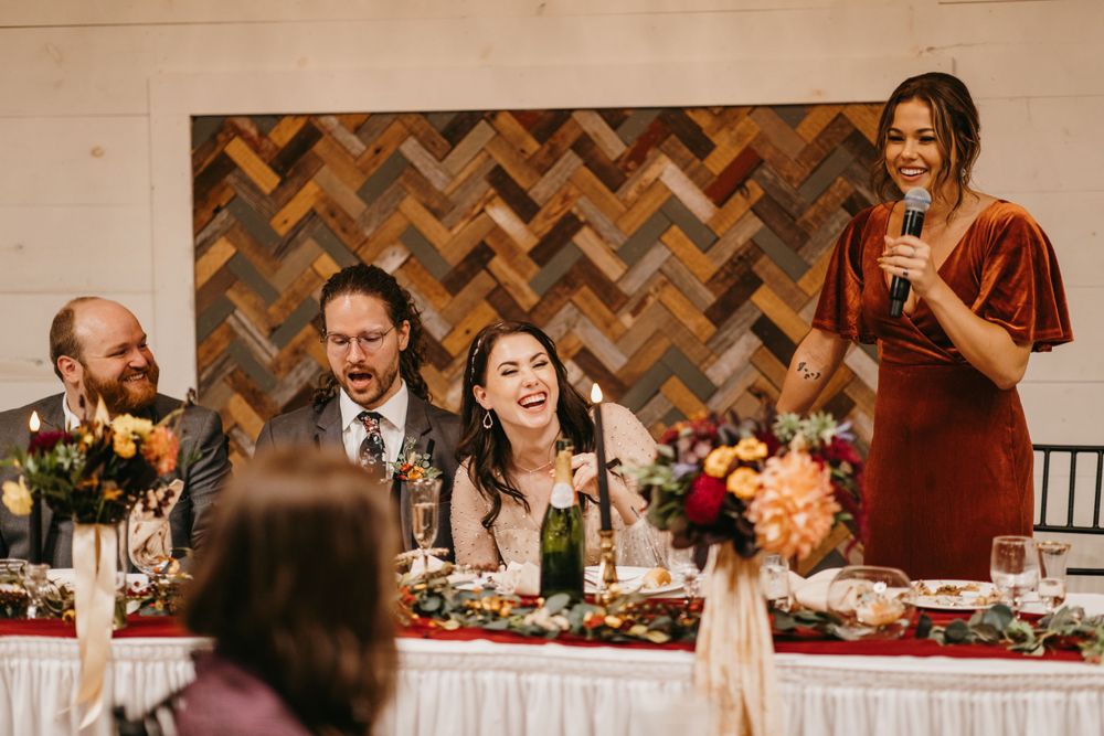 Maid of honor in burnt orange velvet dress delivering a wedding toast as the couple laughs at head table.