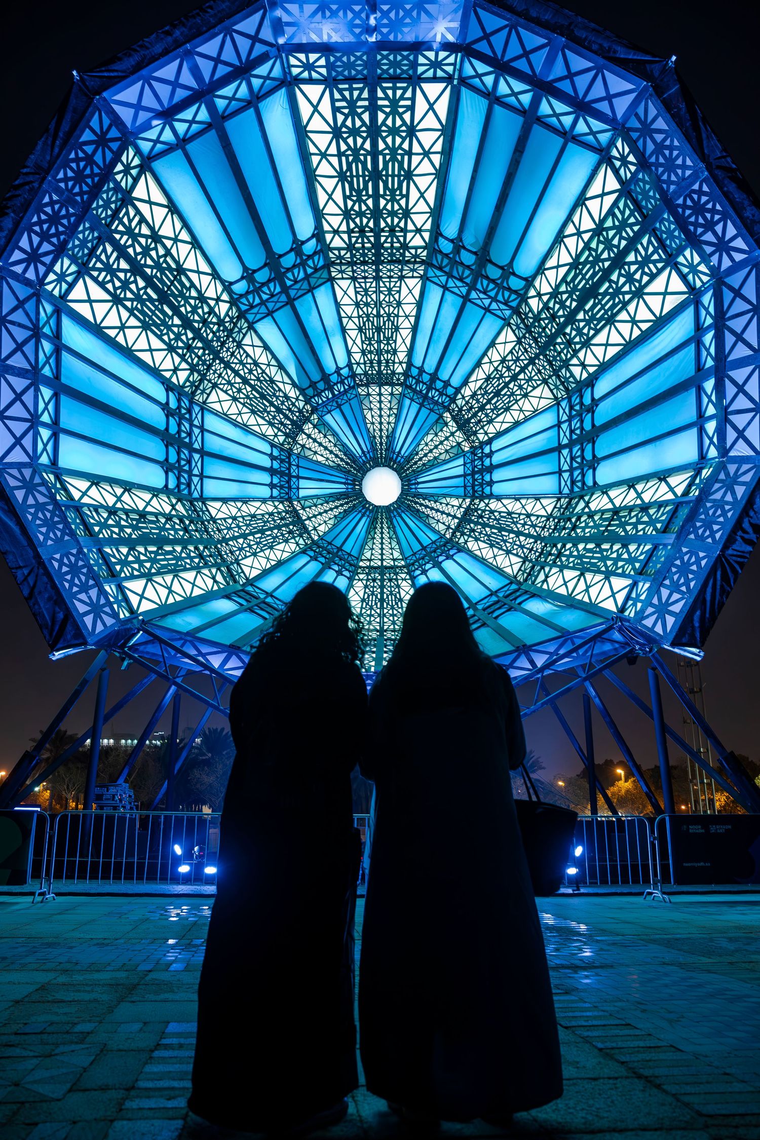 Two silhouetted figures stand before a luminous blue geometric dome structure with intricate lattice patterns.