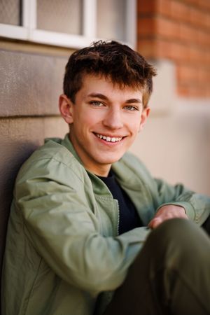 Casual portrait of someone in an olive green jacket leaning against a brick wall outdoors.