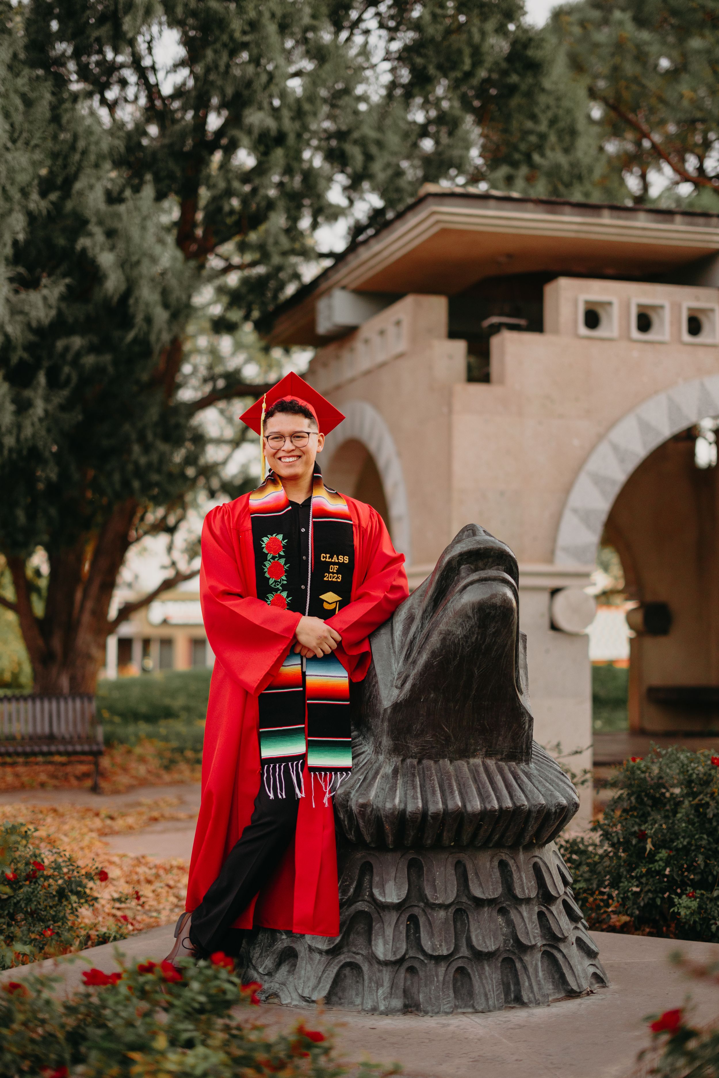 Graduate in red cap and gown posing next to decorative stone knight statue on campus.