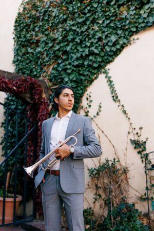 Musician in gray suit holds trumpet against ivy-covered wall.