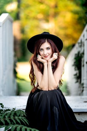 Woman in black dress and hat sits on white bridge in autumn setting.