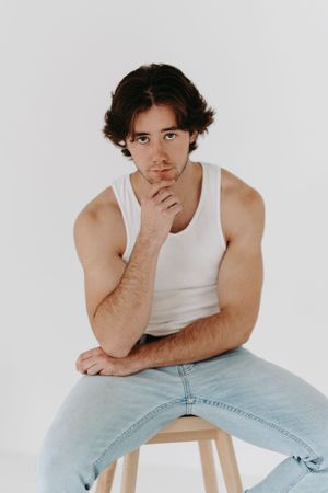 Simple studio portrait of person in white tank top and jeans on wooden stool.