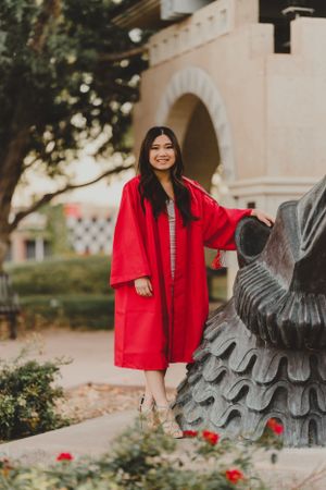 Graduate in bright red graduation gown poses by decorative statue on campus.