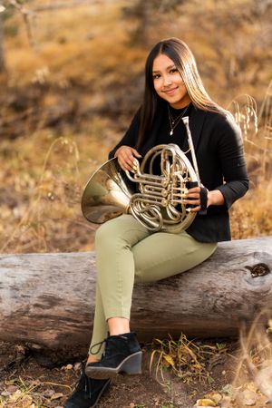 Musician poses with French horn on fallen log in autumn setting.