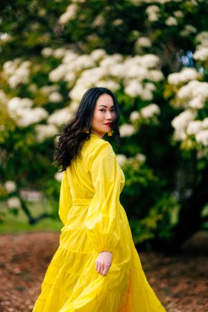 Person in flowing yellow dress poses in garden with blooming white flowers.