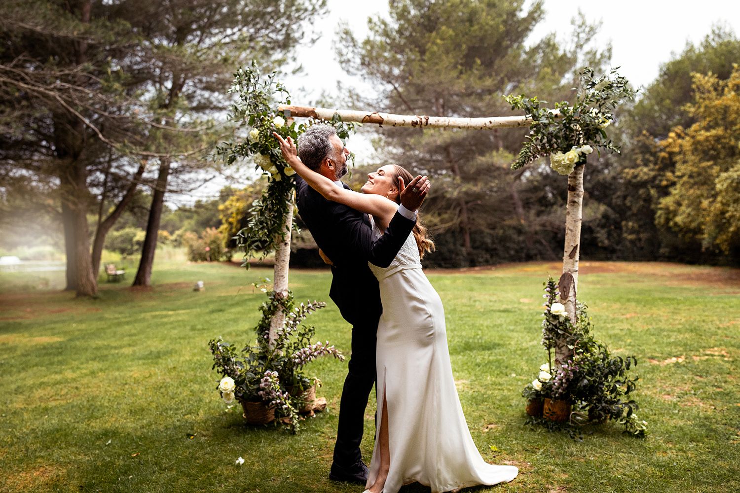 A romantic wedding celebration under a rustic wooden arch decorated with greenery in an outdoor garden setting.
