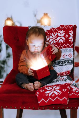 Small child sits on red chair with festive holiday pillows and glowing lights.