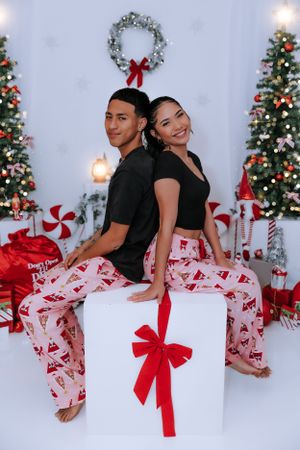 Couple in matching outfits sitting back-to-back on white present prop with Christmas decorations.