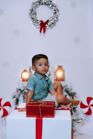 A young child sits on white presents surrounded by holiday decorations and lanterns.