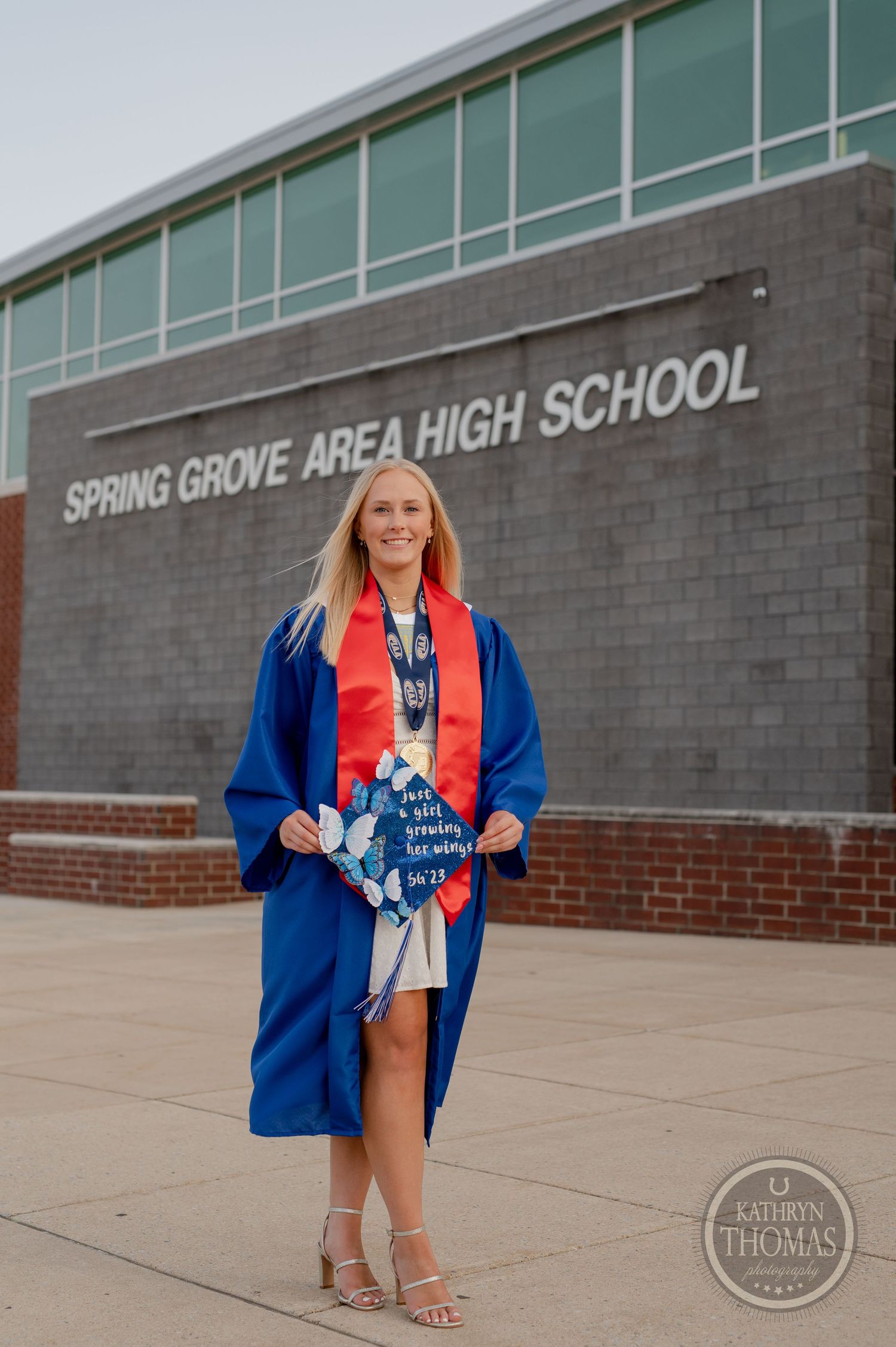 Graduate in blue gown and red stole poses with decorated cap in front of high school.