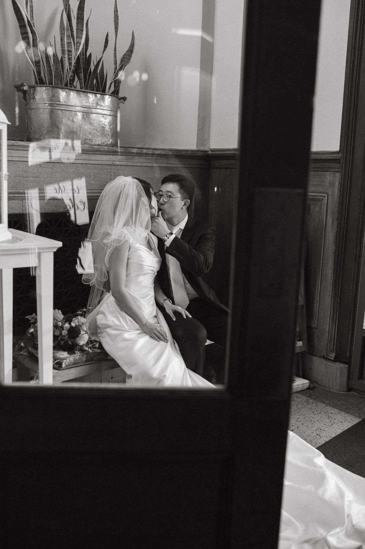 A black and white intimate wedding moment captured through a window with a potted plant.