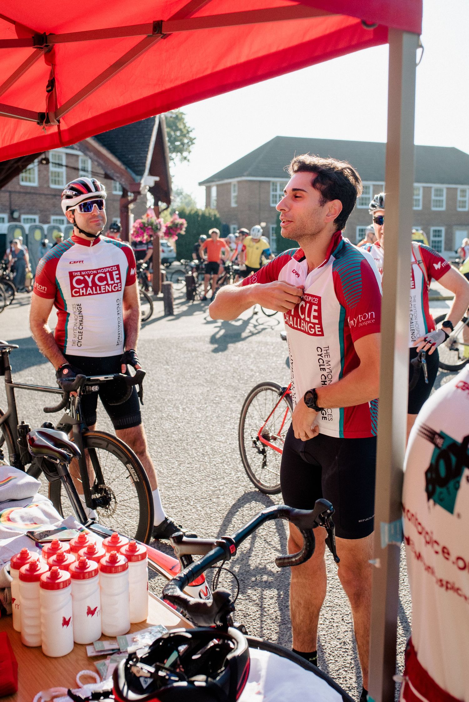 Cyclists at a refreshment station under a red tent during a cycling event.