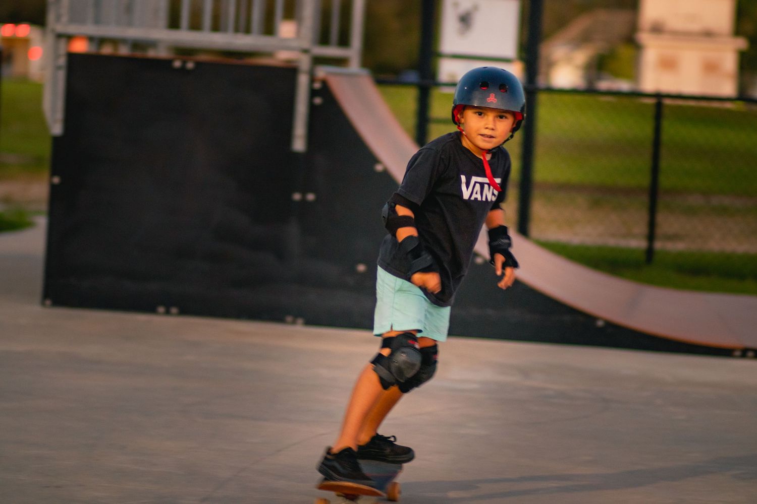Young skateboarder in Vans shirt and blue helmet rides at an outdoor skate park wearing protective knee pads.