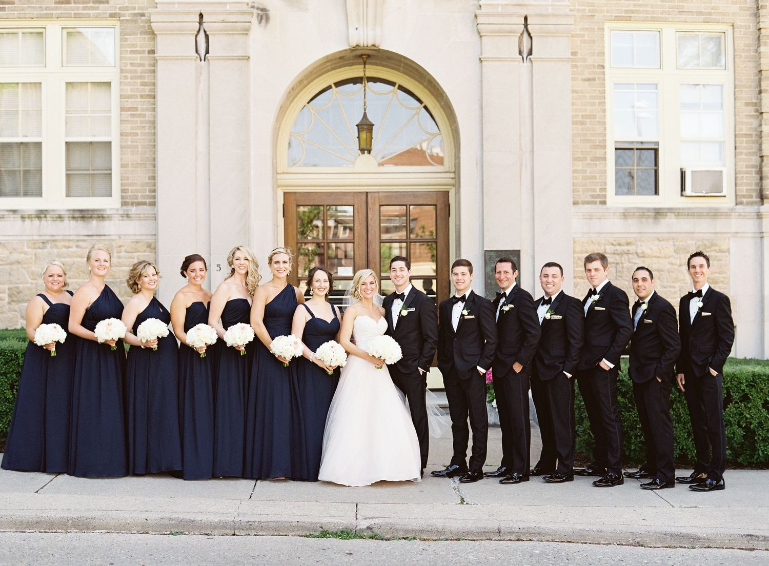 Wedding party in navy and black formal attire poses outside stone building with white bridal bouquets.