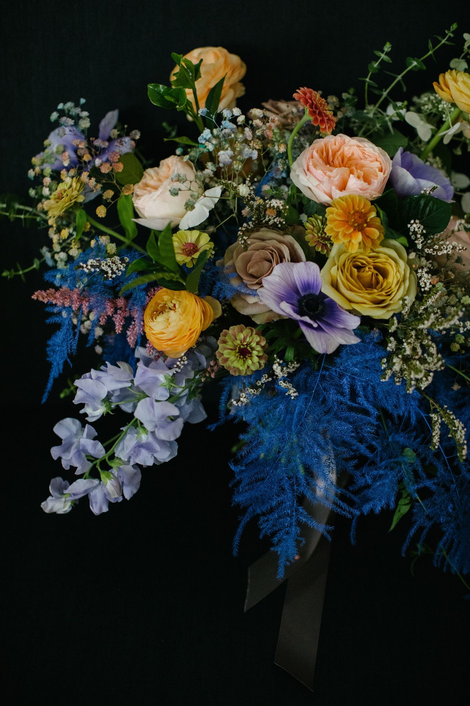 Lush wedding bouquet featuring colorful blooms including peach roses, yellow ranunculus, purple anemones, and blue ferns.