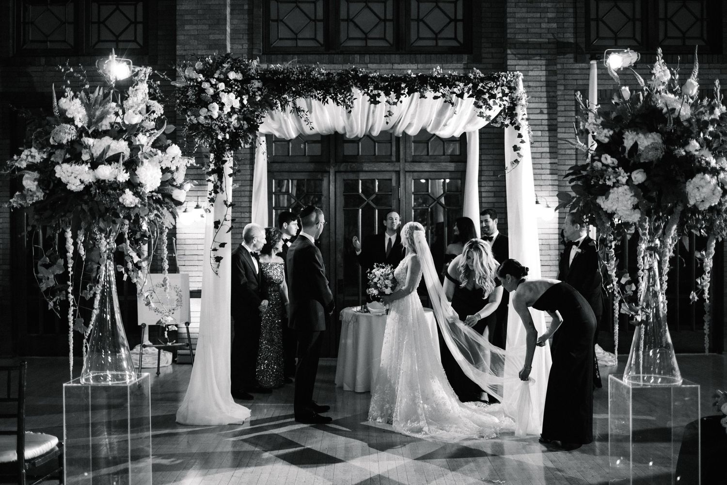 A bride and groom exchange vows under a floral chuppah in an elegant black and white ceremony photo.
