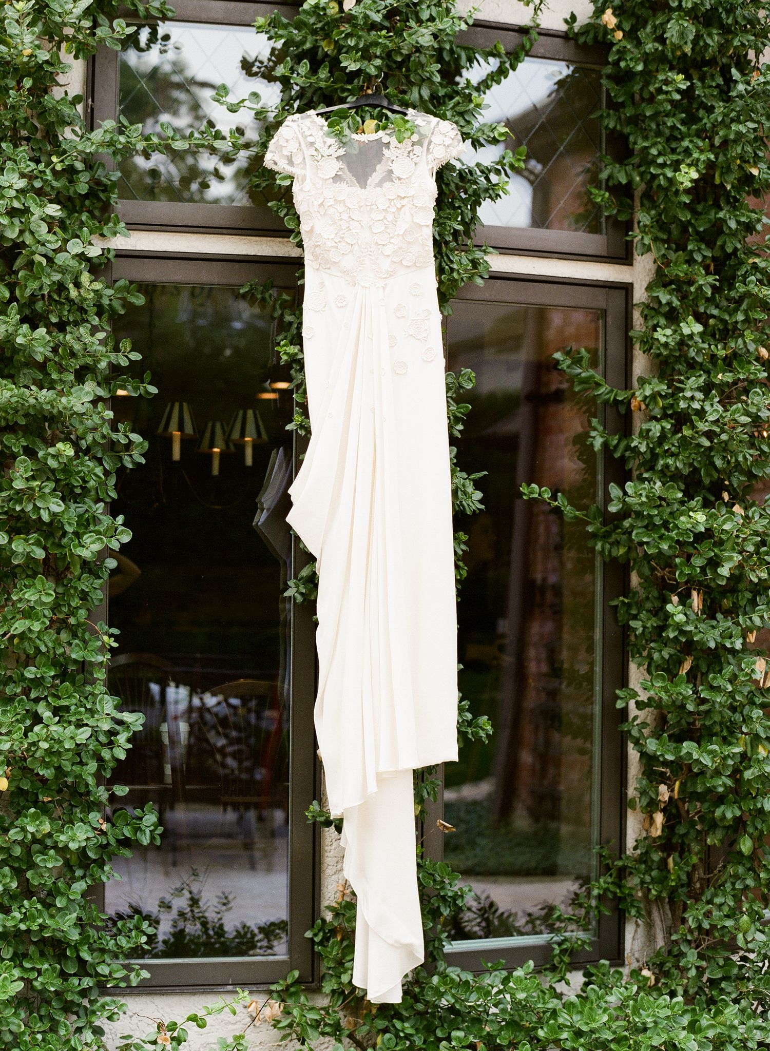 An elegant lace cap-sleeve wedding gown hanging on ivy-covered wall outside a venue.