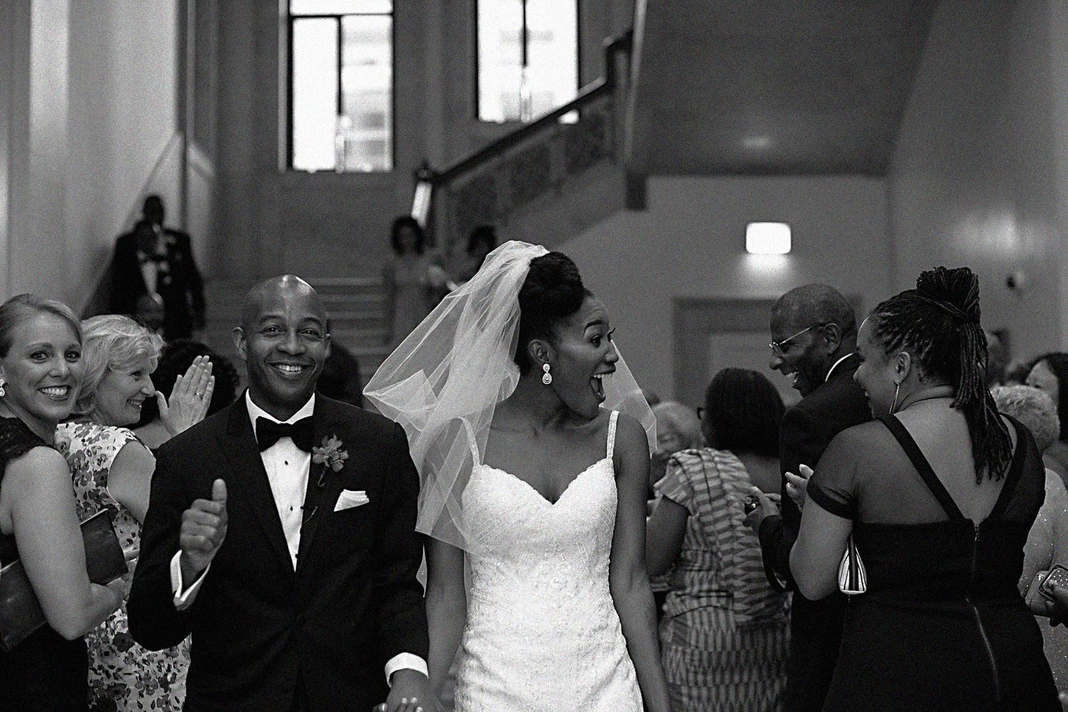 Joyful newlyweds walk down aisle in black and white photo surrounded by cheering wedding guests.