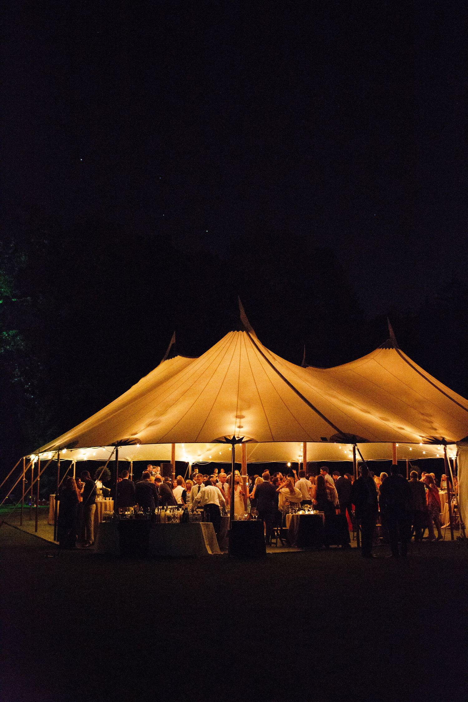 A glowing white wedding reception tent illuminated at night with guests celebrating inside.