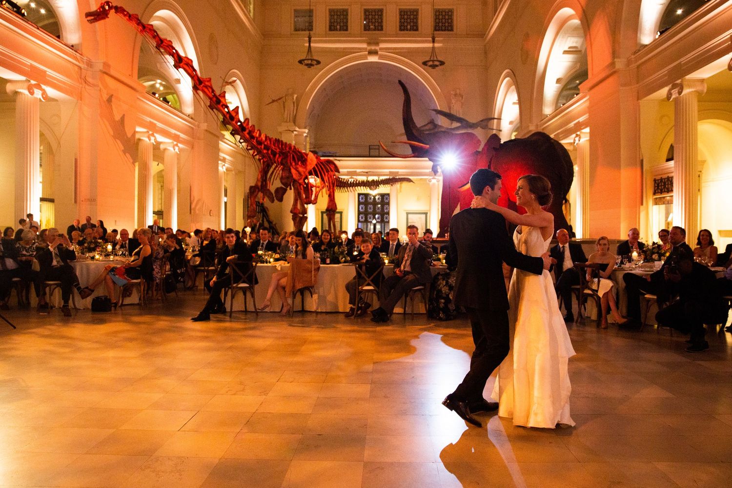 Couple shares first dance at elegant museum wedding reception with dinosaur skeleton display and warm amber lighting.