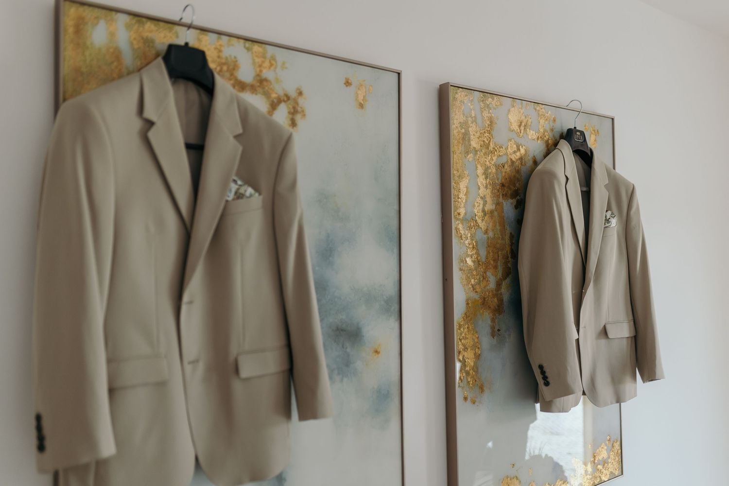 Two matching beige groom suits hanging on wall beside gold abstract paintings.
