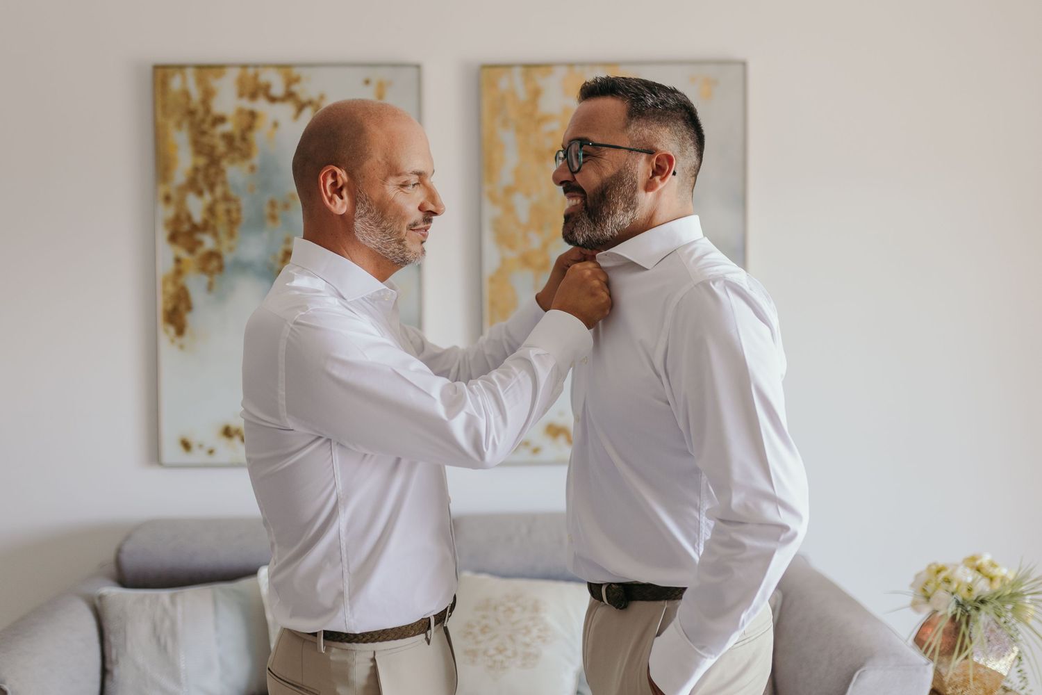 Two grooms helping each other dress in white shirts before wedding ceremony.
